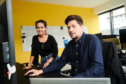 Ein Mitarbeitender sitzt am Schreibtisch im Büro und arbeitet, eine Kollegin steht daneben und blickt auch in den Bildschirm.