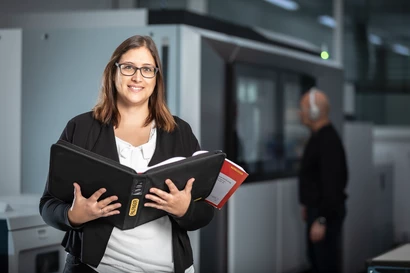 Eine Mitarbeiterin aus dem Controlling hält eine Mappe in ihren Händen und lächelt in die Kamera. Im Hintergrund ist ein Kollege zu sehen, der Kopfhörer trägt.