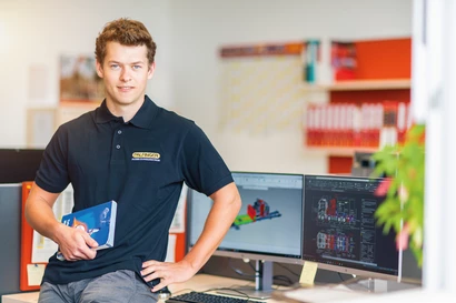 Ein Schüler lehnt am Schreibtisch, hält ein Buch in der Hand und blickt in die Kamera.