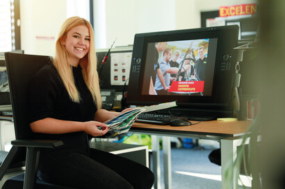 A schoolgirl sits at her desk, holding colour cards in her hand and smiling at the camera.