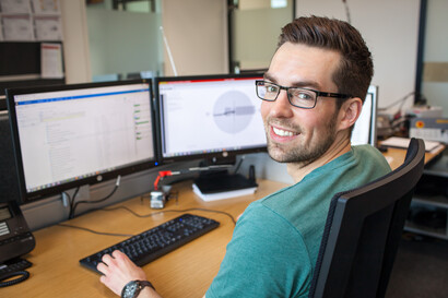Ein Mitarbeitender lächelt in die Kamera. Im Hintergrund sind zwei aktive Bildschirme zu sehen.