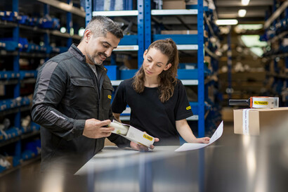 Zwei Mitarbeitende stehen hinter einem Tisch, der Mann öffnet ein Päckchen, das mit einem PALFINGER Logo versehen ist und zeigt der Kollegin den Inhalt. Die Dame hält ein Blatt Papier in der Hand.