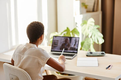 A woman sits in front of her laptop with her back to the camera and takes notes on a piece of paper during a training session.
