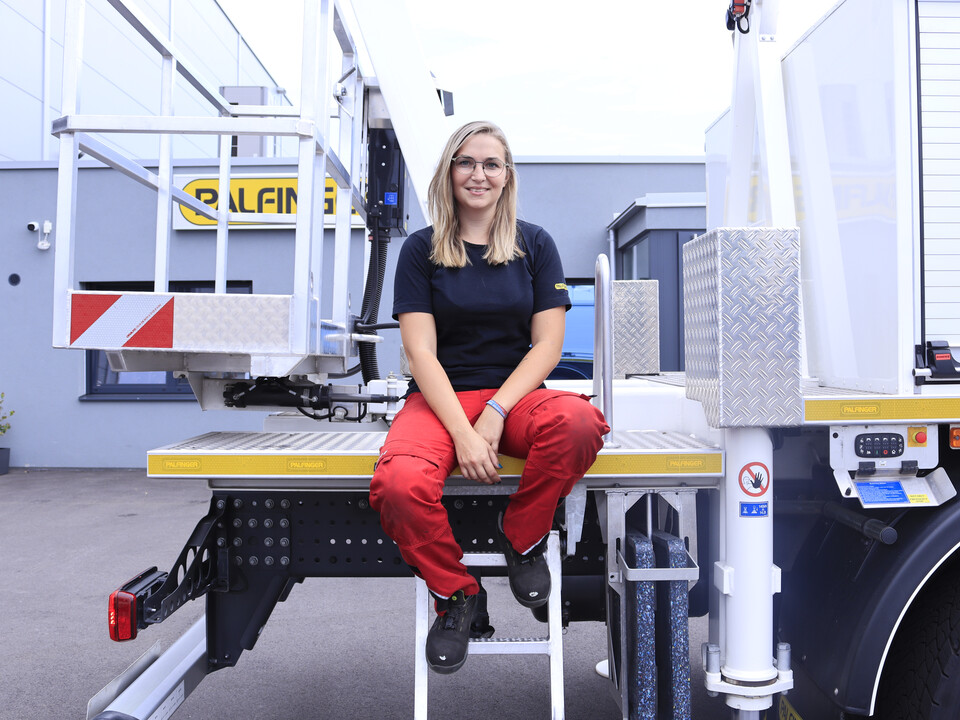 Eine Servicetechnikerin sitzt am hinteren Teil eines LKW mit Hubarbeitsbühne und schaut direkt in die Kamera. Eine Servicetechnikerin sitzt am hinteren Teil eines LKW mit Hubarbeitsbühne und schaut direkt in die Kamera.