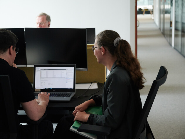 Eine Mitarbeiterin und ein Mitarbeiter sitzen am Schreibtisch mit dem Rücken zur Kamera und schauen in einen Laptop. Im Hintergrund arbeitet ein weiterer Mitarbeiter.