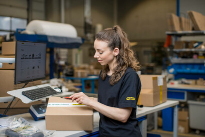 Eine Mitarbeiterin steht vor einem Tisch im Lager und bereitet ein Paket vor.
