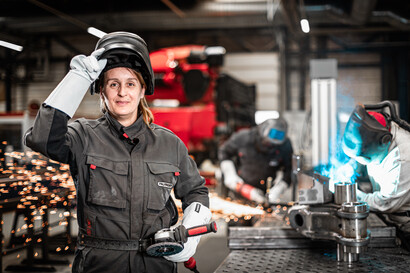 Eine Schweißerin steht im Vordergrund und lächelt in die Kamera. Im Hintergrund arbeiten zwei weitere Schweißer.