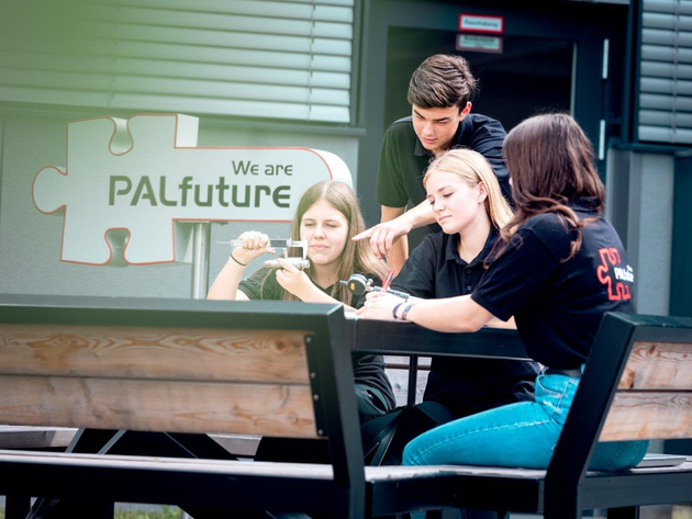 Vier Jugendliche sitzen an einem Tisch und arbeiten an einer Aufgabe. Im Hintergrund  sieht man das Palfuture Logo.