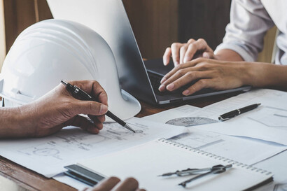 Two employees are working at a desk. One is working on a laptop, the other is writing something on a piece of paper. A hard hat is lying next to the laptop.
