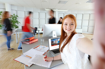 Eine Mitarbeiterin steht vor ihrem Laptop und im Hintergrund sieht man ein paar verschwommene Personen gehenitzen am Schreibtisch mit dem Rücken zur Kamera und schauen in einen Laptop. Im Hintergrund arbeitet ein weiterer Mitarbeiter.