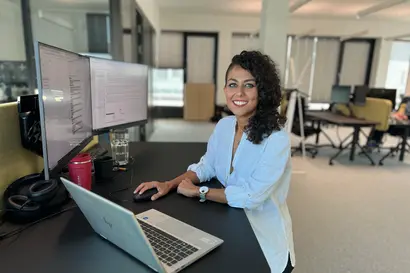 An IT employee stands at her desk and smiles at the camera. | © A PALFINGER employee can be seen working in an office on two screens and a laptop. Another workstation can be seen in the background