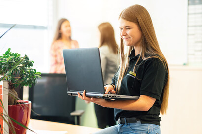 A trainee is working on her laptop. In the background, 2 colleagues are chatting with each other.