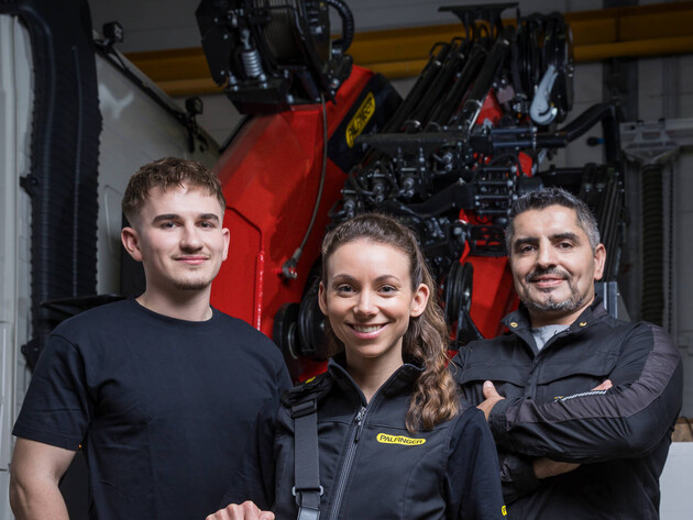 Drei Mitarbeitende stehen vor einem Ladekran und blicken in die Kamera. Eine Mitarbeiterin hält die Steuerung des Kranes in der Hand.